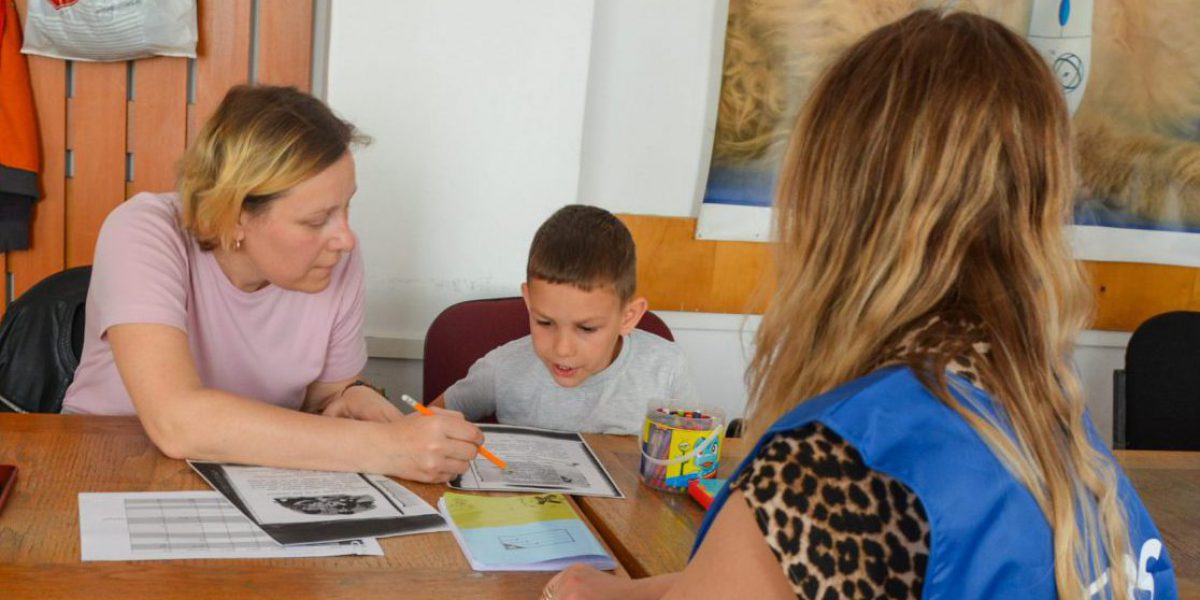 Two women and a child sitting at a table. One woman is helping the child write, and the other woman has her back facing the camera.