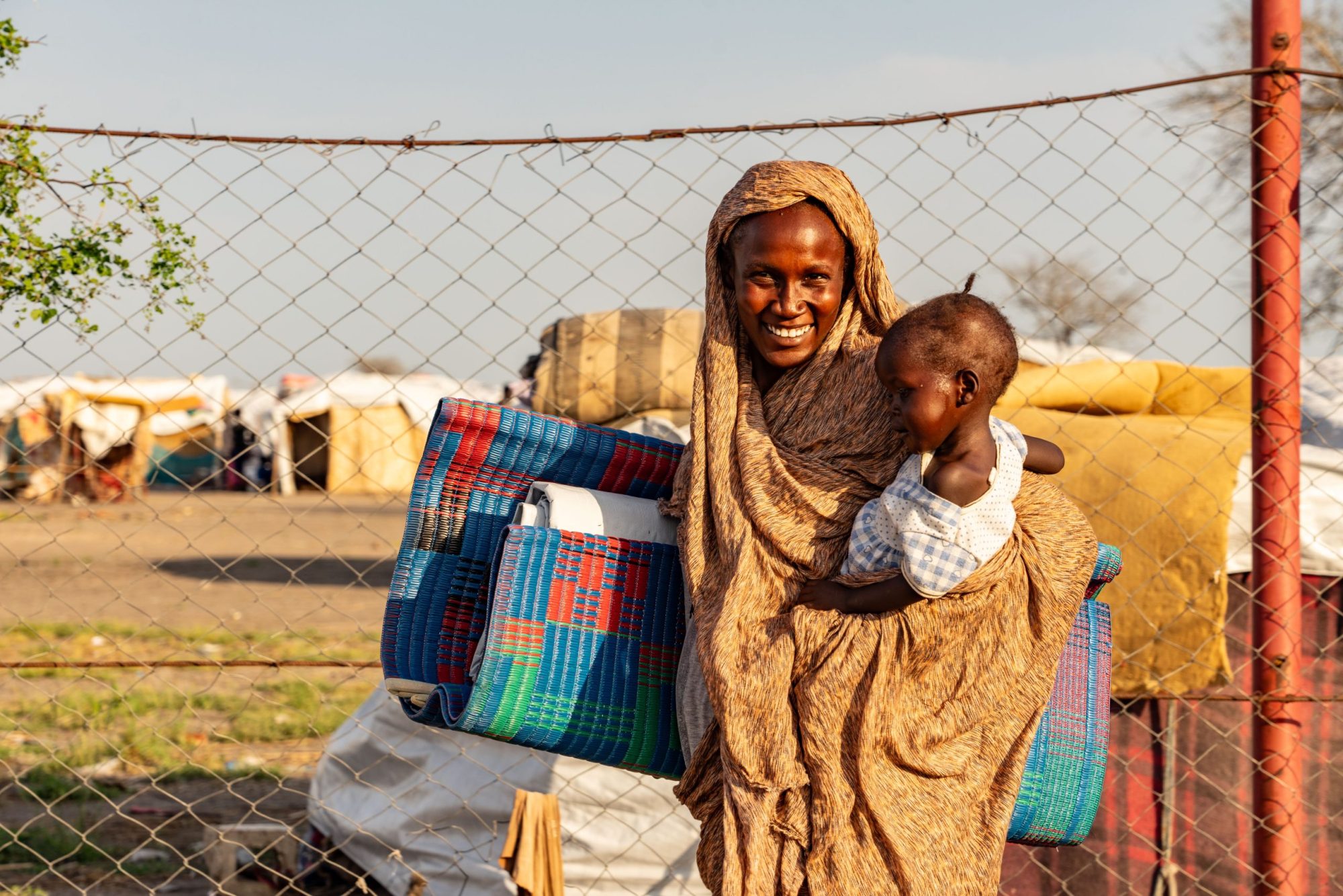 A mother and a baby standing outside of a barbed wire fence smiling.